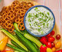 An overhead shot of a bowl of Seven Onion Dip Spice & Easy on a board with pretzels and fresh vegetables.